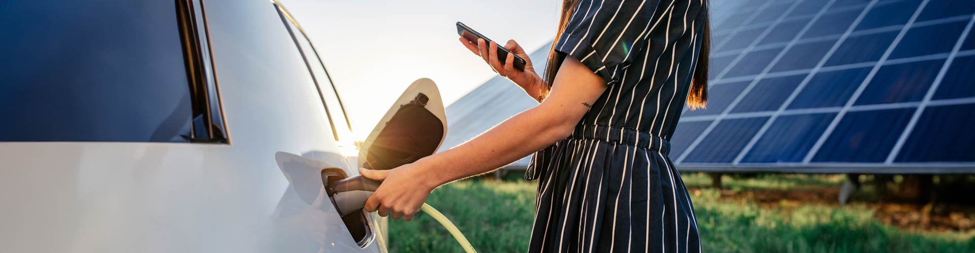 A woman holds a phone while she charges an EV in front of a row of solar panels