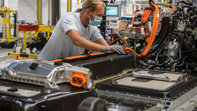 A man with a long blond ponytail leans over a long black box and wires on an assembly line in a warehouse.