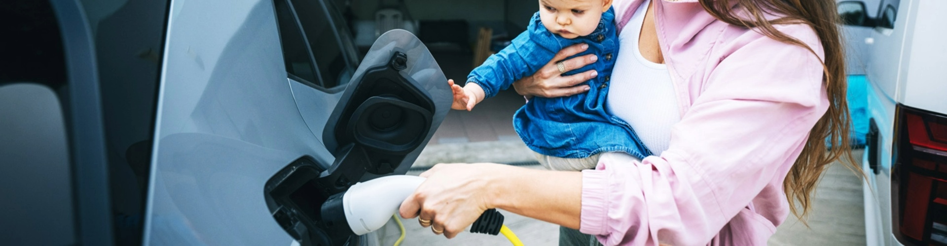 A mother holding an EV charging cable in one hand, plugging into her car, while holding her baby on her hip with her other hand.