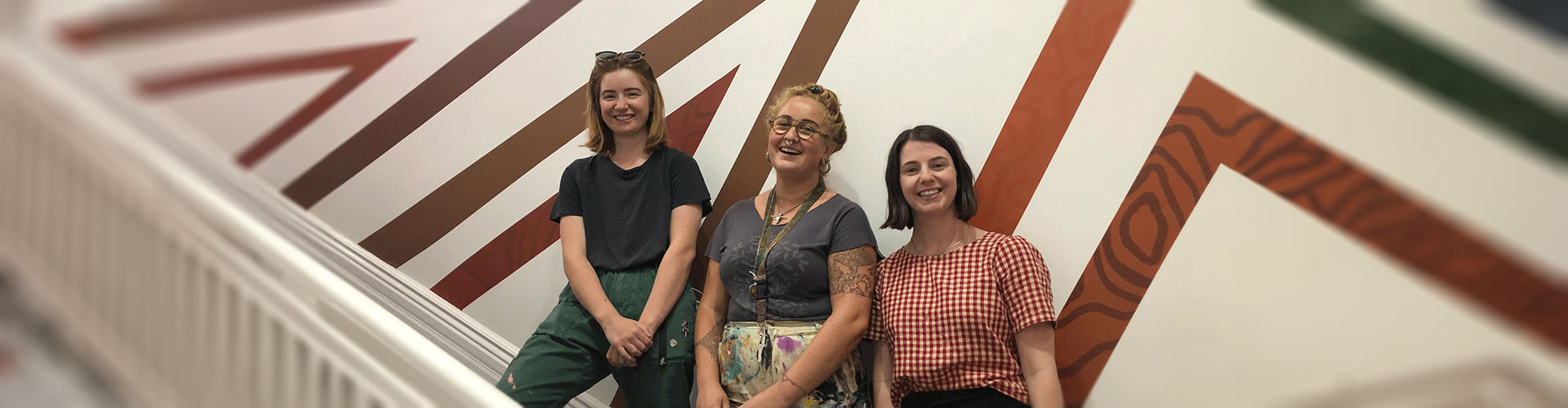 Three women stand on stairs in front of a wall with large geometric brown designs