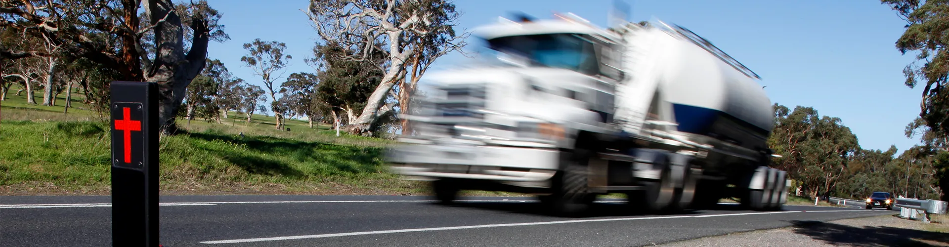 Heavy transport truck speeding past a road fatality marker (a black post with a red cross) on an Australian highway