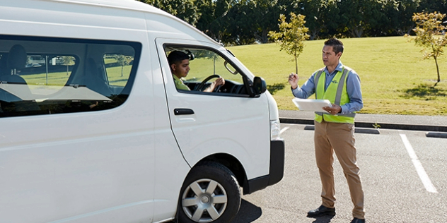 A man with a clipboard giving instructions to a man driving a white vehicle.