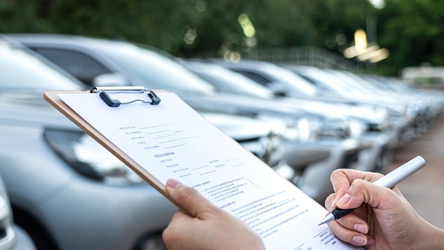 Someone goes through a checklist on a clipboard, while looking at a row of cars 