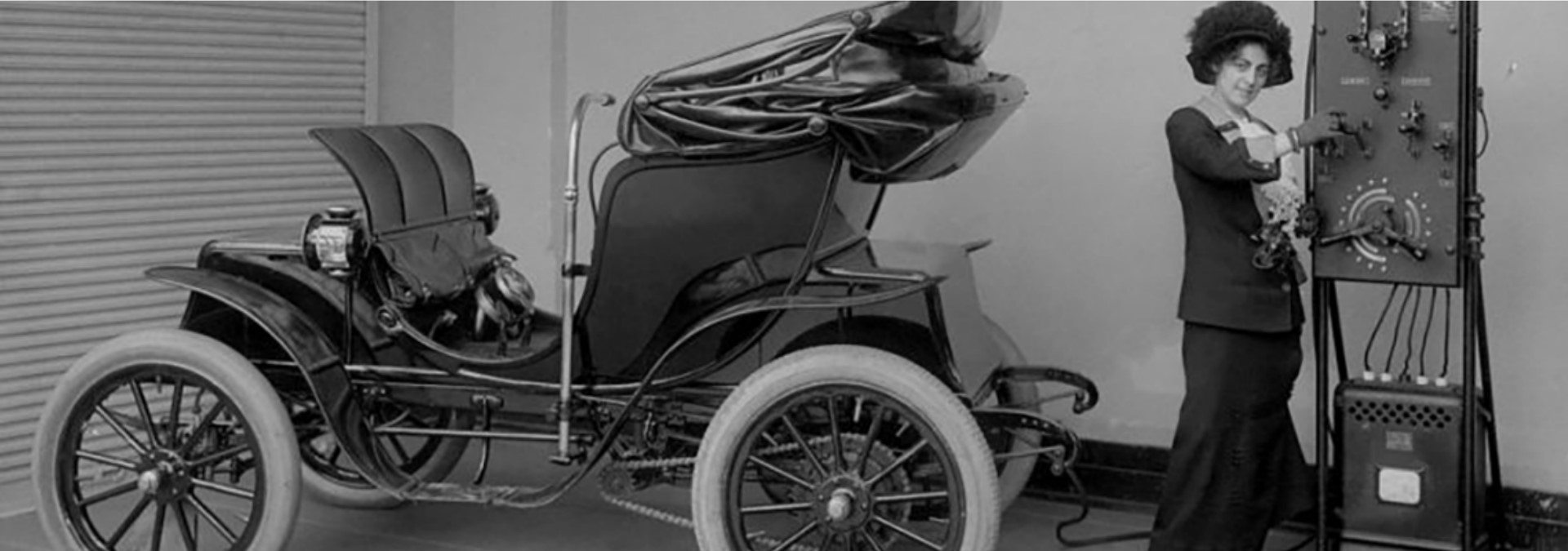 woman-charging-historical-ev-1920x675 A black and white photo of a model-T like car, with a woman in a hat, dress and gloves standing behind it, connecting a cord to an electrical panel.