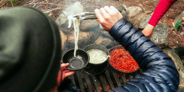 A person puts pasta in their camp bowl sitting next to a campfire with a pan of sauce and pot of pasta
