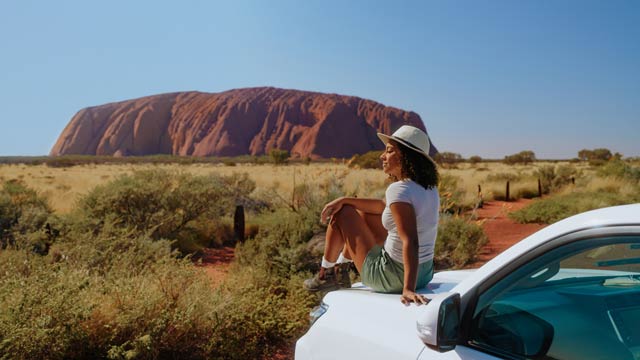 A woman sits on a car bonnet with Uluru in the distance