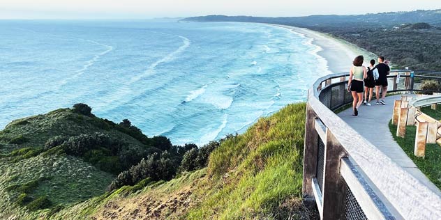 People walk on path past view over Tallow Beach from Byron Bay Lighthouse 