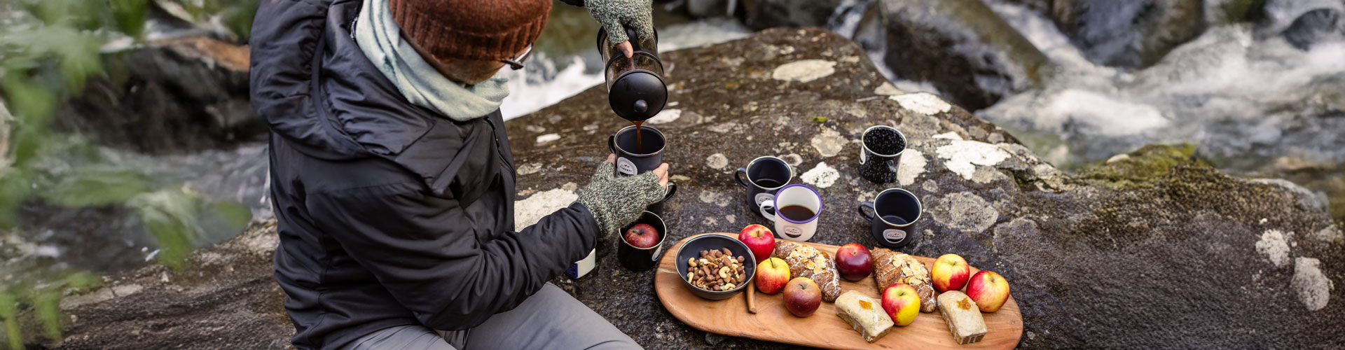 A man pours a hot drink sitting next to a rocky creek with a wooden board of apples and snacks