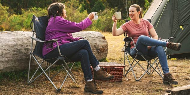 Two women sitting in camp chairs holding mugs outside a tent 