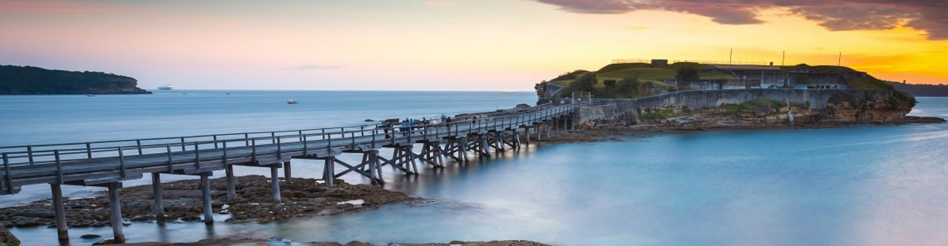 bare-island-botany-bay-nsw-1920x500 A wooden walking bridge out to a small green island with historic fort buildings, lit from behind by sunset.