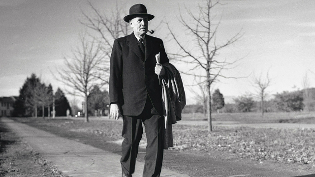 A black and white photo of former Prime Minister Ben Chifley in his sixties, wearing a hat and smoking a pipe, walking down a sidewalk.