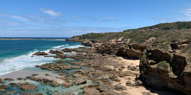  A beach with rounded, grassy cliffs and flat rocks dotting the sand leading out to clear, turquoise water. 