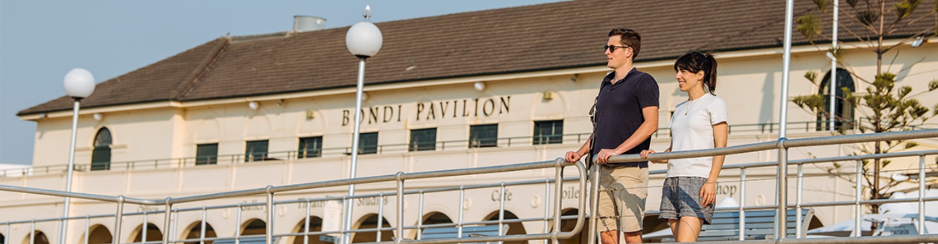 bondi-pavilion-building-1920x500 A man and woman lean against a metal railing along a boardwalk lined with vintage, round street lamps, on a cloudy day. Behind them is a long off-white building with the 'Bondi Pavilion' written in upper case New Roman font above the top row of windows.