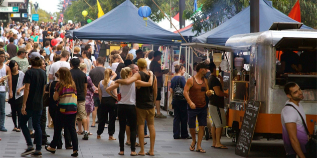 A sunny, bustling, city market with marquees and food trucks. 