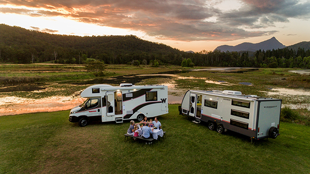 Group of people sitting at a table together besides a winnebago caravan and motorhome lakeside