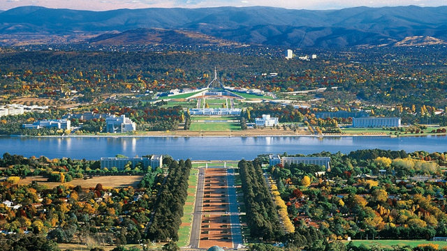 A panoramic view of Canberra city, with parliament house in the centre in front of a lake, with flat treed plains and a mountain range behind it.