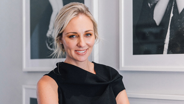 canna has blond, tied-back hair and has a warm smile. She wears an asymmetrical black top and her head and shoulders are visible