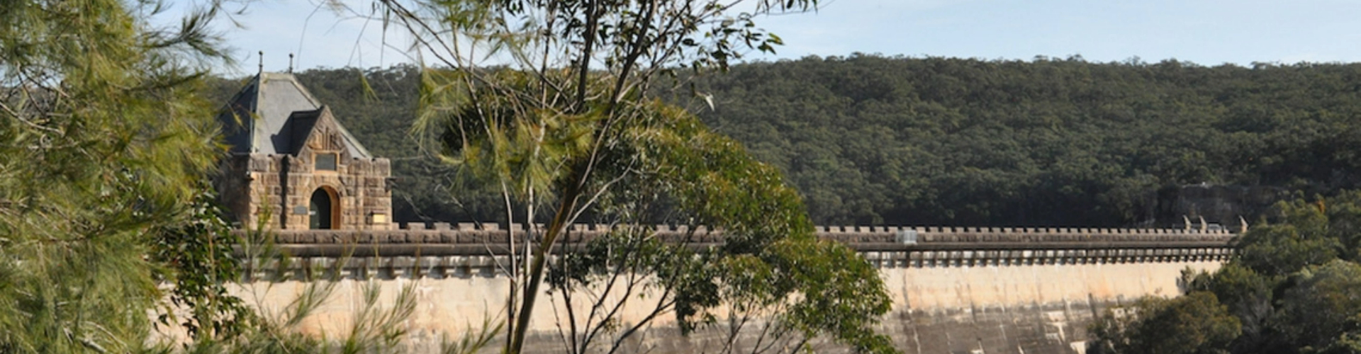 cataract-dam-nsw-1920x500 A maintenance building that looks like a stone house at one end of the top of a dam, surrounded by a forest.