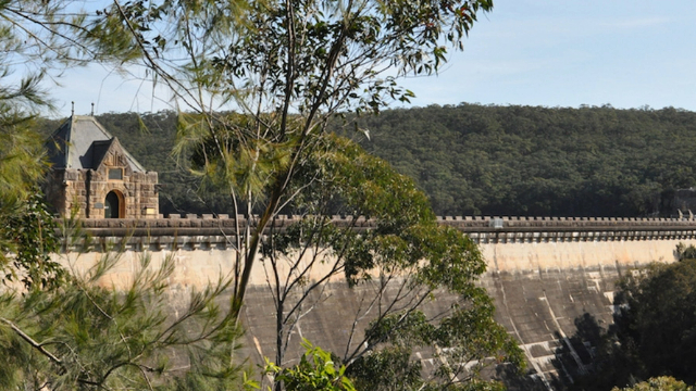 A maintenance building that looks like a stone house at one end of the top of a dam, surrounded by a forest.