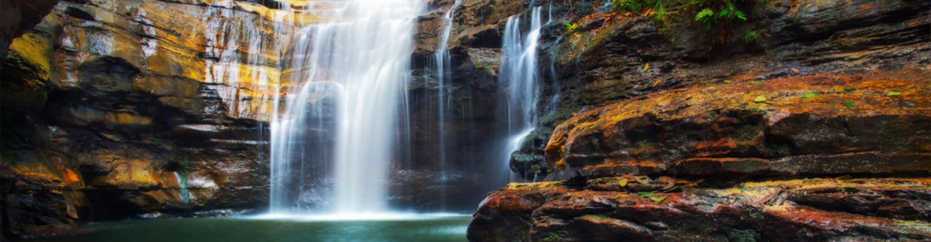 cave-waterfall-nsw-1920x500 A waterfall lit with sunlight as it pours into a cave over rocks and into a dark pool of green, surrounded by mottled red, brown and yellow cave walls.