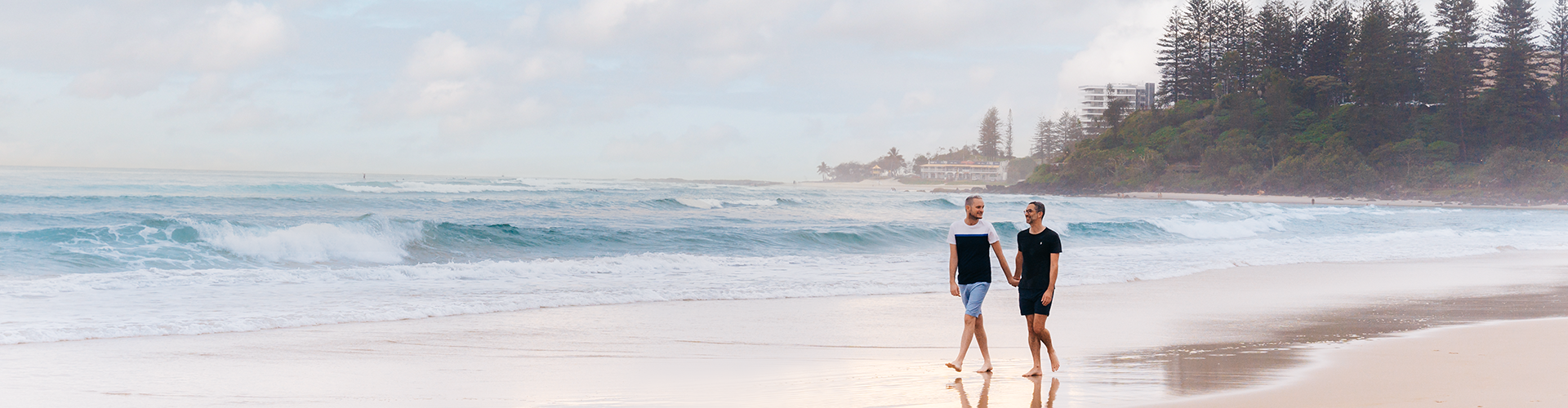 Two men walk along the shore of Coolangatta Beach, holding hands