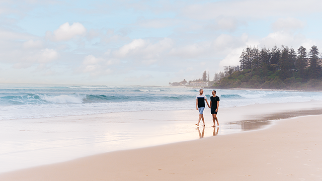 Two men walk along the shore of Coolangatta Beach, holding hands