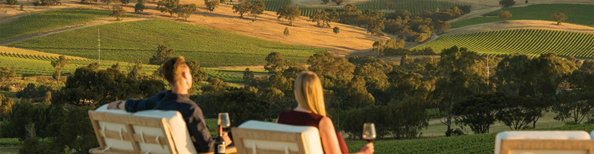 couple-at-winery-barossa-sa-1920x500 A man and a woman sit in chairs on a hill drinking wine at sunset, overlooking green hills and vineyards.