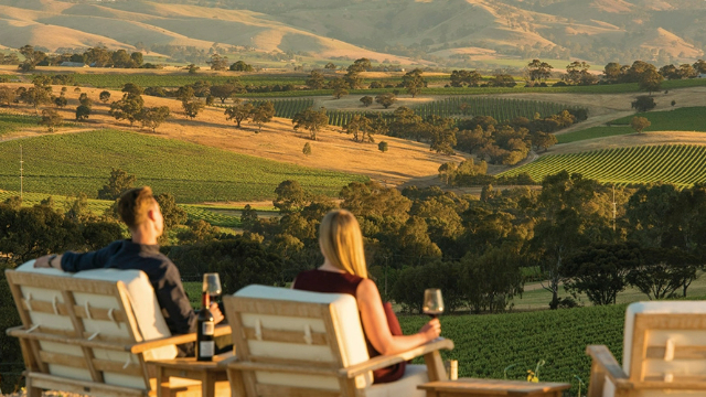 A man and a woman sit in chairs on a hill drinking wine at sunset, overlooking green hills and vineyards.