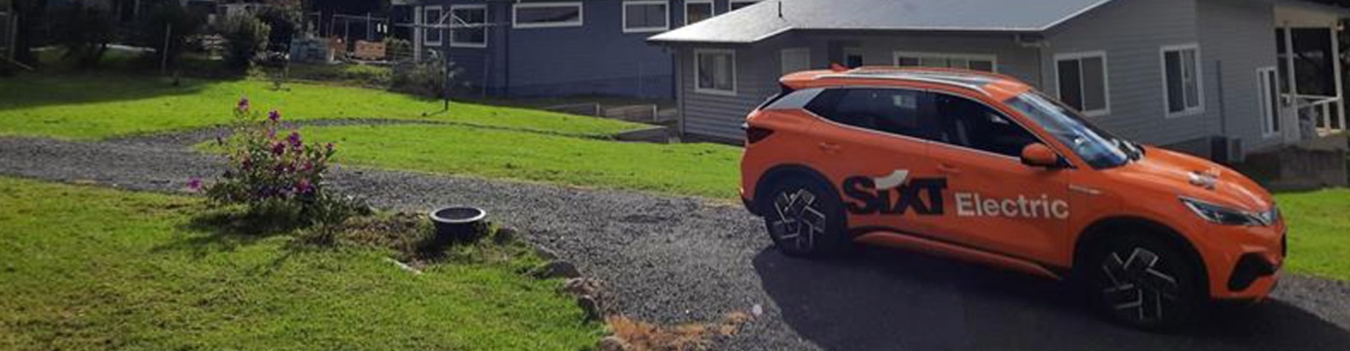 EV Sydney to Lake Conjola road trip orange EV rental car branded SIXT parked outside a grey weatherboard house