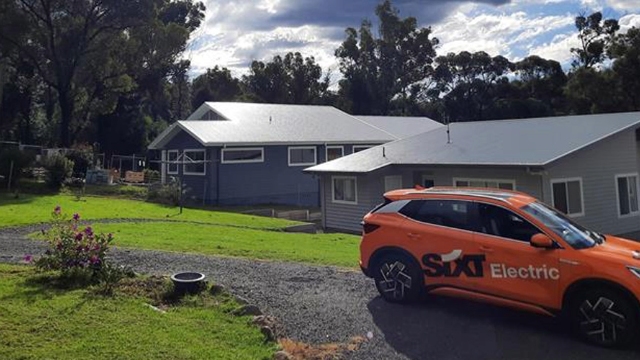 orange EV rental car branded SIXT parked outside a grey weatherboard house