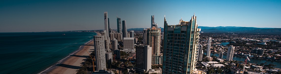 gold-coast-skyscraper-view-1920x500 A city with several skyscrapers along a beach, as seen from another skyscraper window, framed by the ocean and mountains on the horizon.