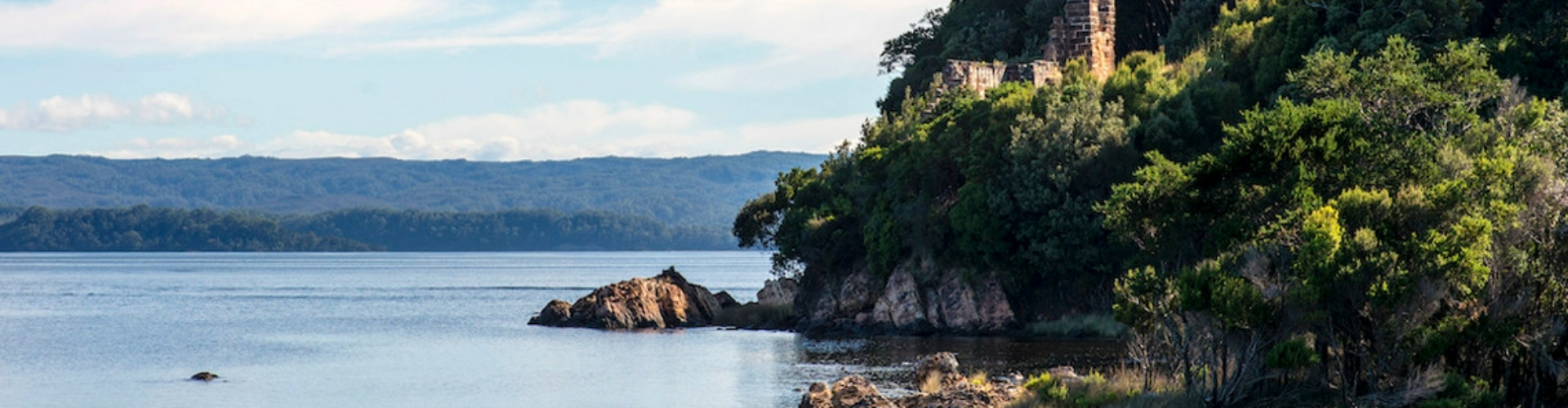 gordon-river-cruise-tas-1920x500 A rocky shore, covered with trees with the remnants of a small brick building on the top of the rocks along the Gordon River.