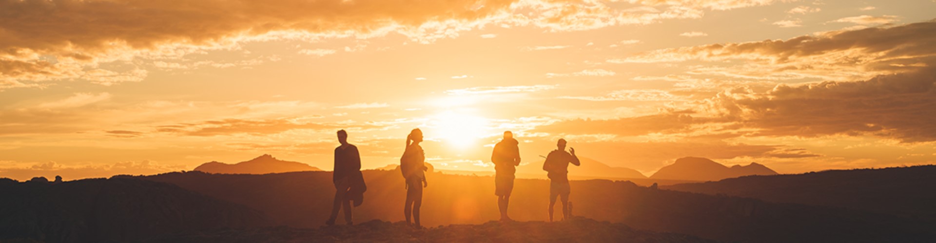 group-in-front-of-sunset-1920x500 Three men and a woman silhouetted by a yellow sunset over hills and mountains in the distance.