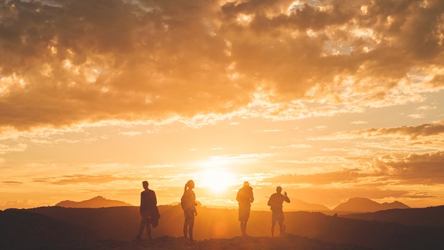 Three men and a woman silhouetted by a yellow sunset over hills and mountains in the distance.
