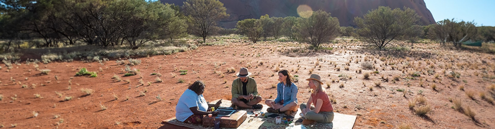 group-sits-at-foot-of-uluru-nt-1920x500 An indigenous woman and three people sit on blanket on the red ground near the base of Uluru rock on a sunny day.