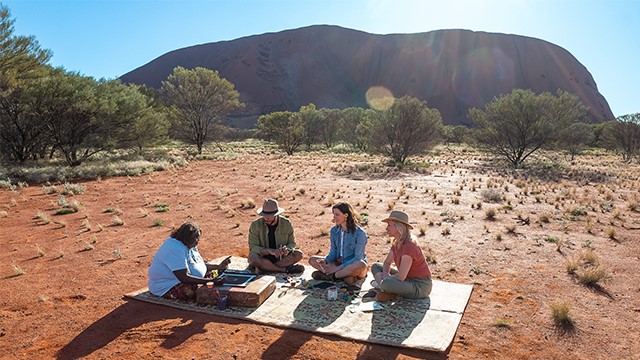 An indigenous woman and three people sit on blanket on the red ground near the base of Uluru rock on a sunny day.  