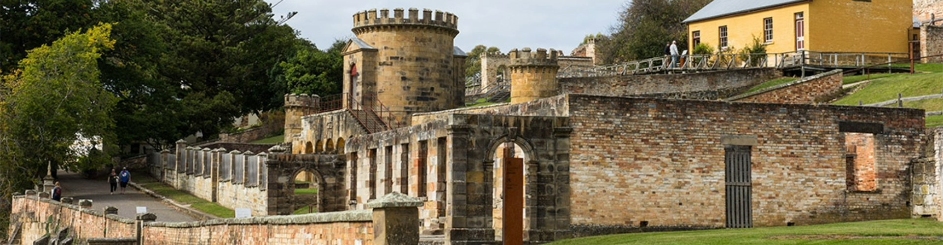 guard-tower-port-arthur-tas-1920x500 A castle like turret with low set brick walls around it, on a grassy hill framed by trees.