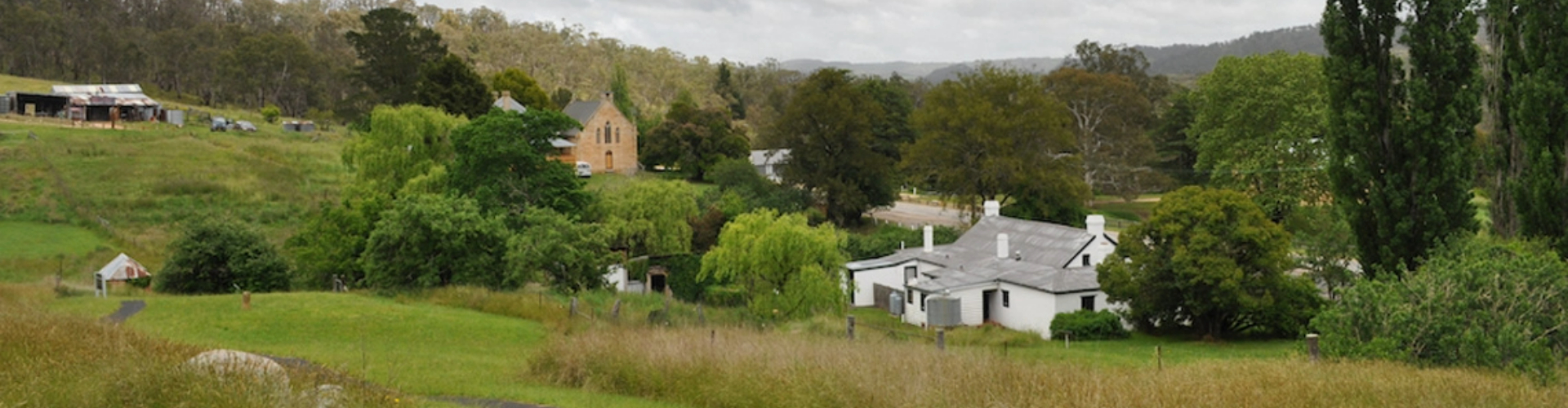 hartley-village-nsw-1920x500 A worn path through lush green grass leads to a few homes and a church in a treed valley.
