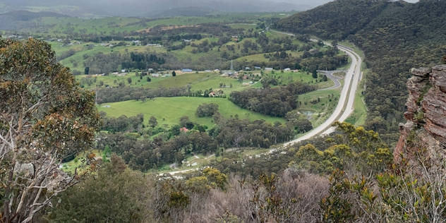  The view from a layered rock cliff looking down over treed hills to a rich green valley, road and village below.