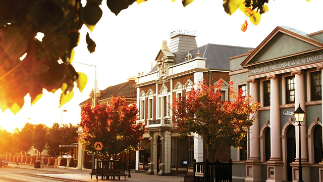 Historic Victorian and colonial style buildings along a street, lit by a flash of orange sunset through tree leaves.