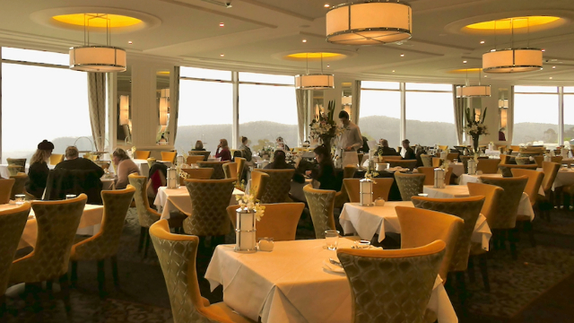 A few patrons in a warm lit, fine dining room with tall windows all around where a silhouette of a mountain can be seen.