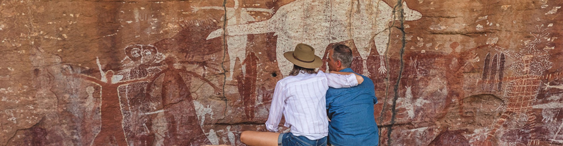 indigenous-art-on-rock-qld-1920x500 A couple sitting together on red rocks in front of a rock wall covered in ancient Indigenous art.
