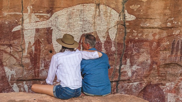 A couple sitting together on red rocks in front of a rock wall covered in ancient Indigenous art.