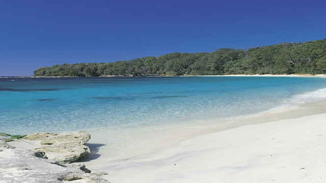 A white sand beach with bright blue water in a bay surrounded by coral like rocks and green trees.