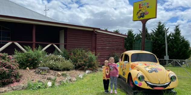 A girl and smaller boy stand by a yellow VW Beetle mounted on grass, painted with a smile and eyes as headlights.
