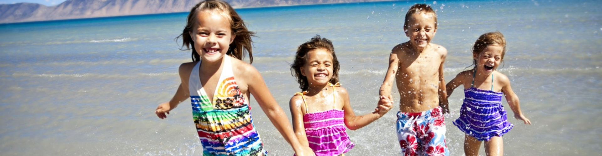 kids-running-together-on-beach-1920x500 Three girls and a boy in swimsuits laugh and hold hands as they run along a sunny beach.