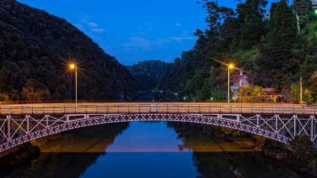A curved metal bridge over a tree covered river valley, lit by two streetlights at dusk. 