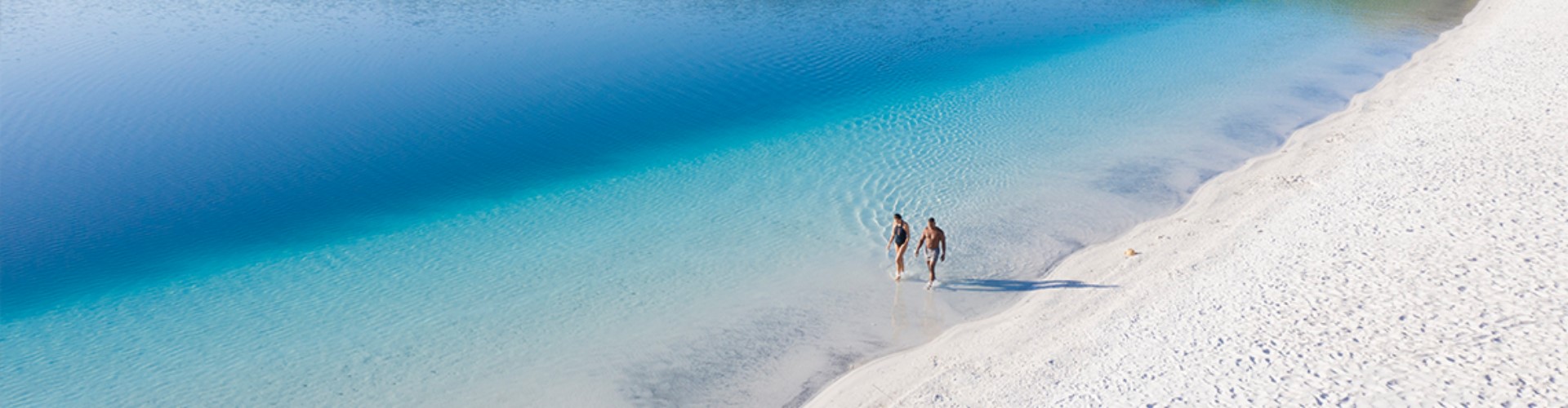 lake-mckenzie-kgari-qld-1920x500 Two people walking along a totally white sand beach leading into clear then deep blue lake waters.