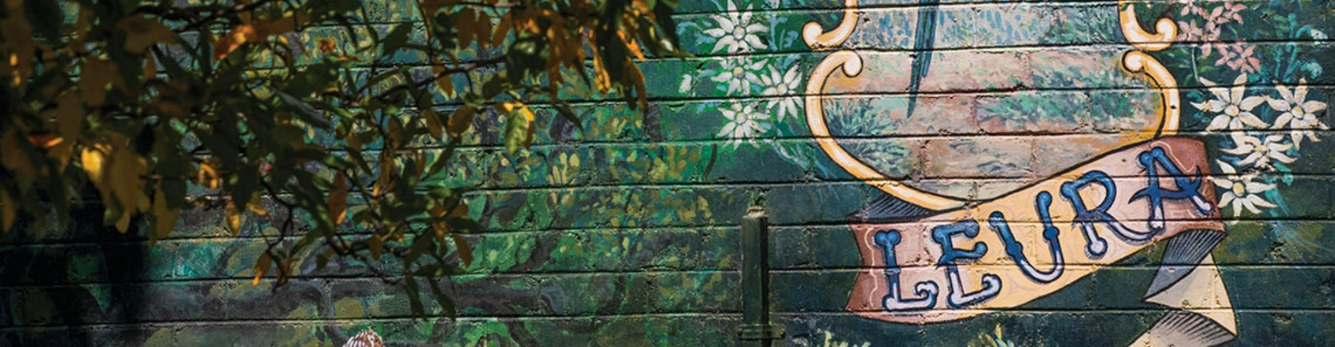 leura-street-art-nsw-1920x500 Street art on a wooden shop wall that shows the word Leura, written on a banner and surrounded by flowers.