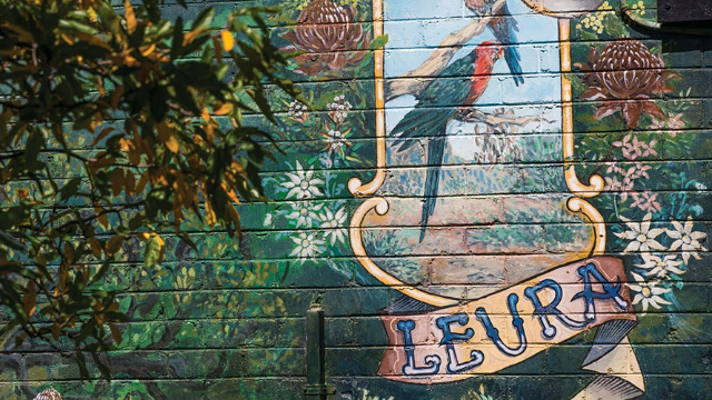 Street art on a wooden shop wall that shows the word Leura, written on a banner and surrounded by flowers.